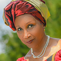 A close-up portrait of an older Black woman, dressed in a red and gold head wrap, adorned with a pearl necklace and a red blouse — Ora Gibson Richards